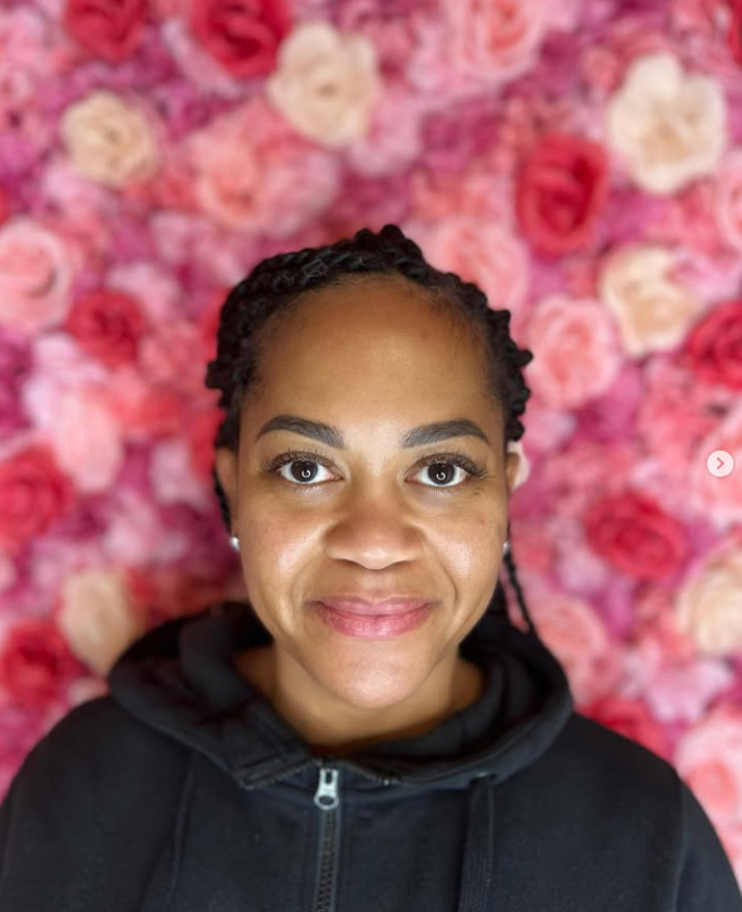 A woman is smiling in front of a wall of pink roses