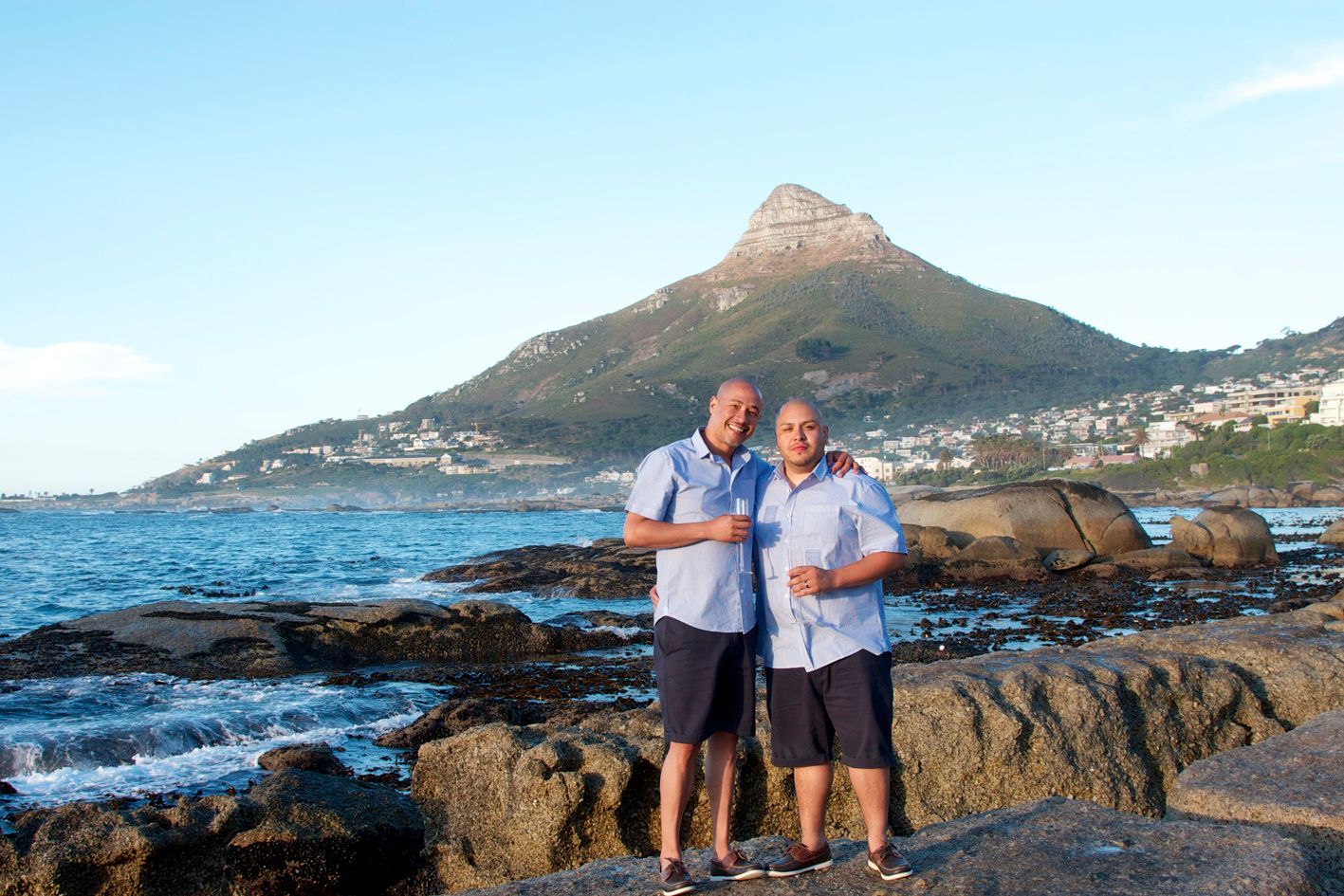 Two men are standing next to each other on a rocky beach with a mountain in the background.