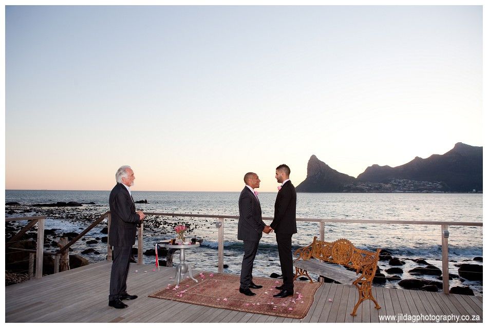 Two men are holding hands while standing on a deck overlooking the ocean.