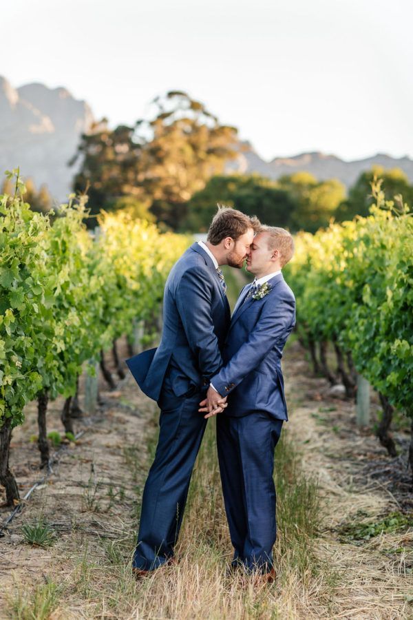 Two men in suits are kissing in a vineyard on their wedding day.