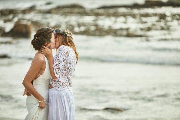 Two women in wedding dresses are kissing on the beach.