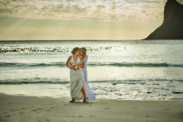 Two women in white dresses are standing on a beach.