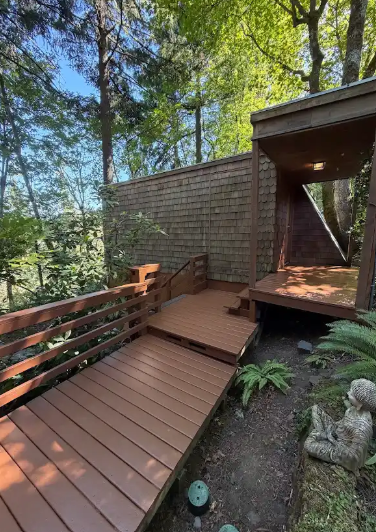 Wooden ramp and deck beside a brick house, surrounded by tall trees and forest greenery.
