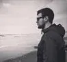 Person in a dark jacket stands by the water, looking toward the horizon on a cloudy day.