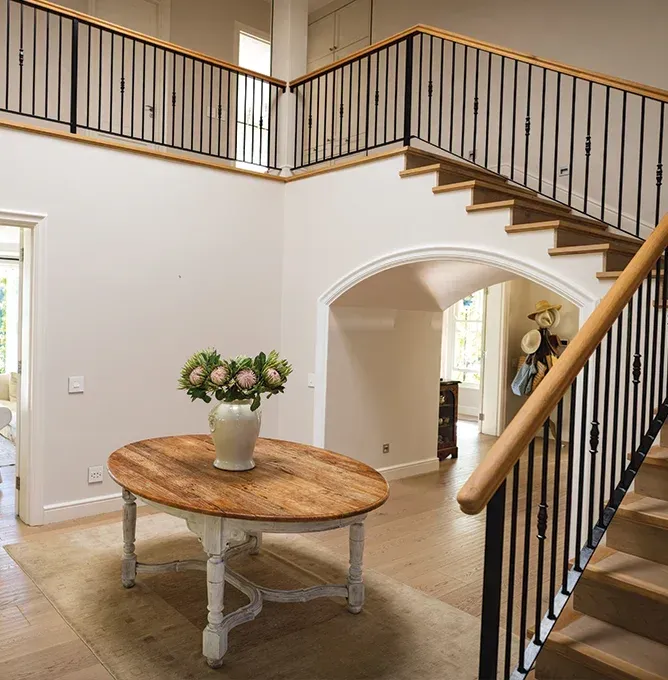 Bright foyer with staircase, round wooden table, and flowers beneath an upstairs balcony.