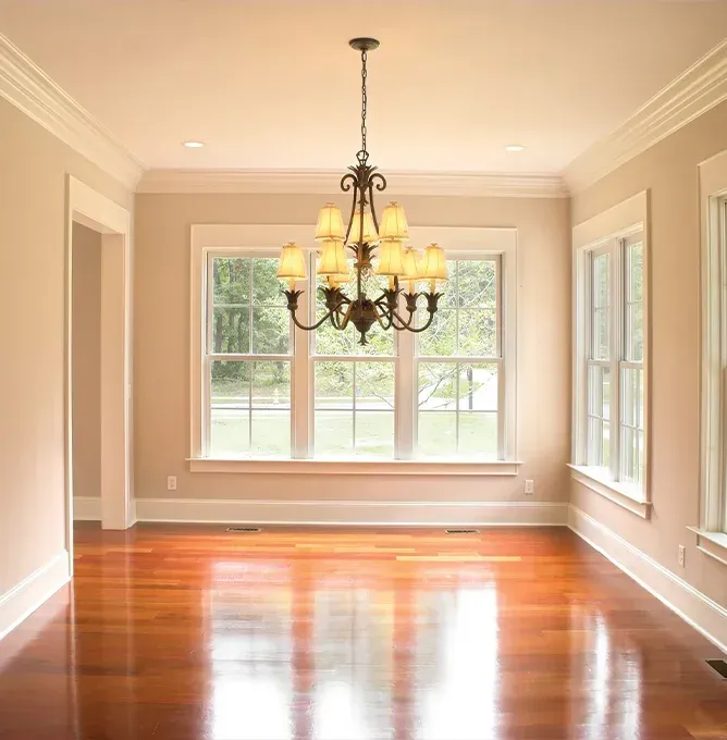 Empty sunlit dining room with polished hardwood floors, beige walls, large windows, and a hanging chandelier