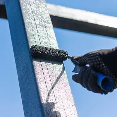 Gloved hand painting a blue metal beam with a roller against the sky