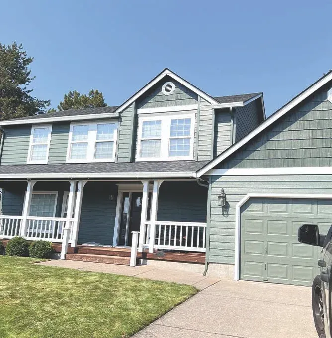 Suburban two-story gray house with white trim, front porch, driveway, and a green garage door under a blue sky