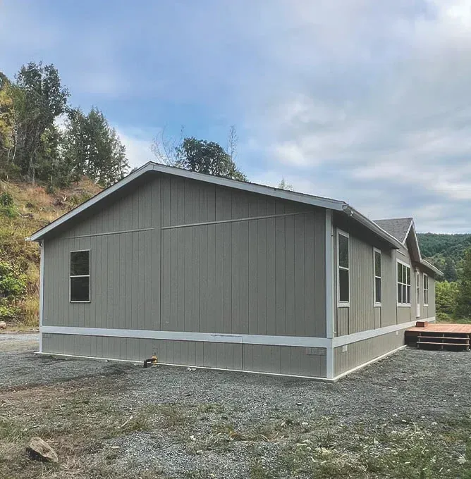 Gray single-story house with white trim on a gravel lot, set against a wooded hillside.