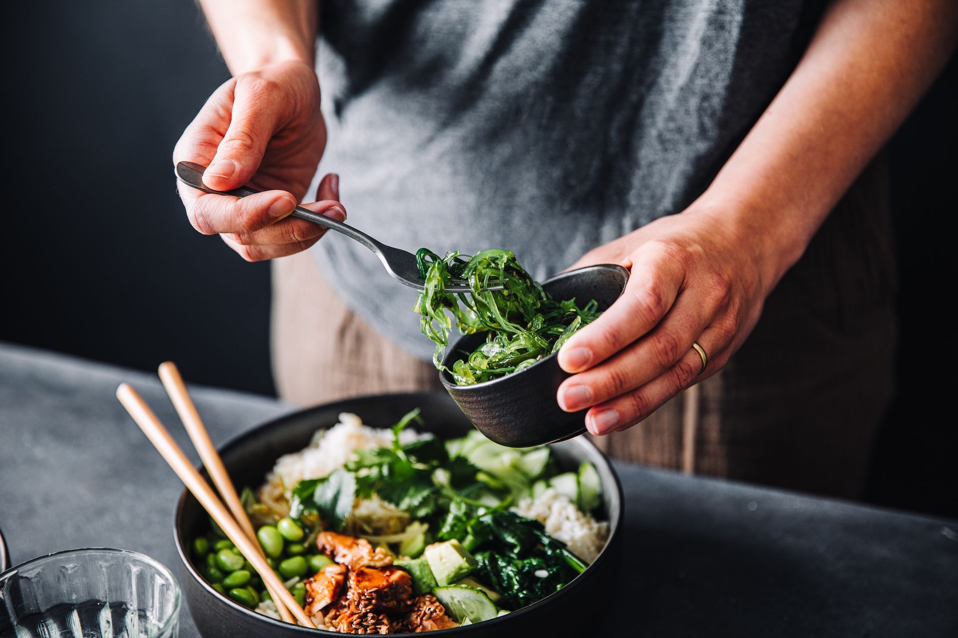A person is adding greens to a bowl of food with chopsticks.
