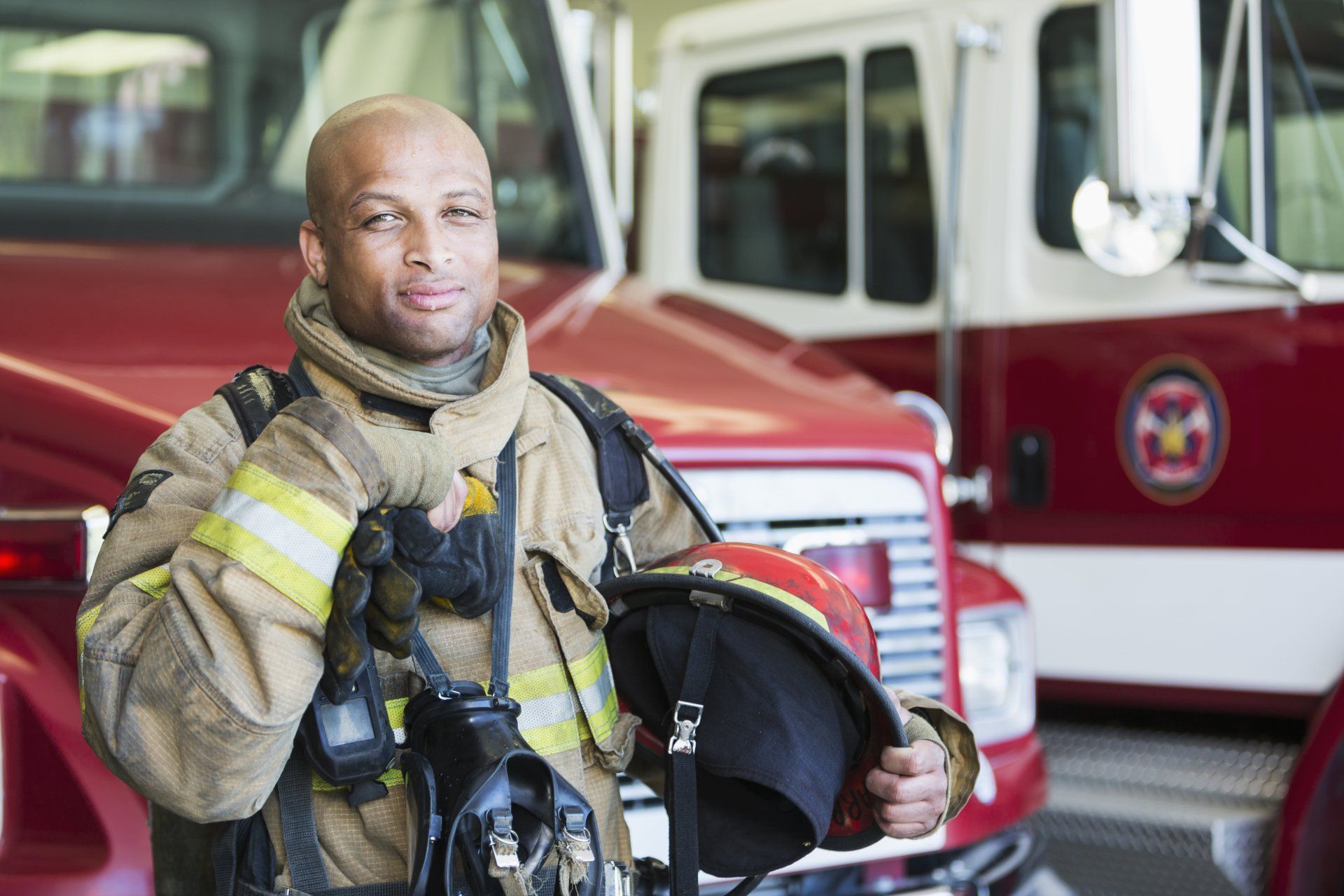 Firefighter in protective gear holding helmet, standing in front of firetrucks.