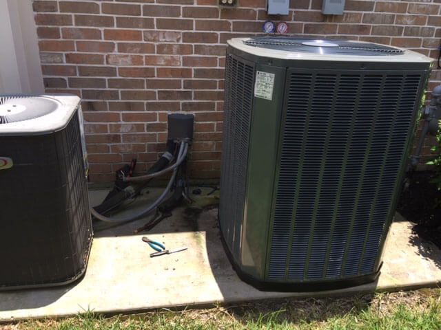 Two air conditioners are sitting next to each other on a sidewalk in front of a brick building.