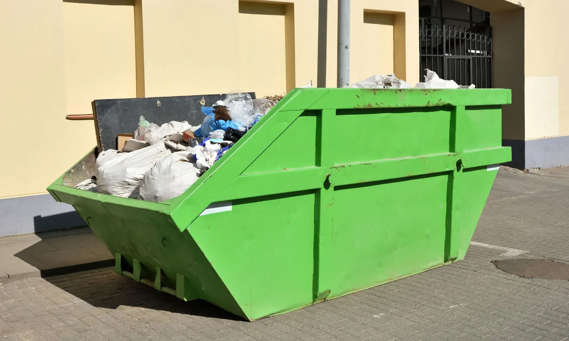 Bright green metal dumpster filled with trash on a street beside a yellow building
