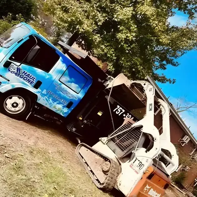 Blue and white trucks parked beside a house under trees, including a compact construction vehicle with a bucket