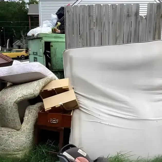 Outdoor cluttered yard with stacked mattresses, boxes, a green dumpster, and a gray fence behind.