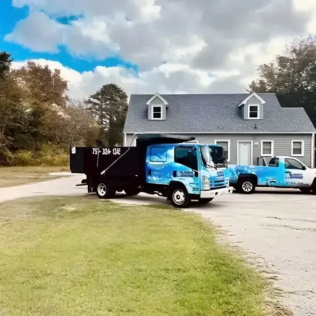 Blue moving trucks parked outside a gray house on a grassy driveway under a cloudy sky