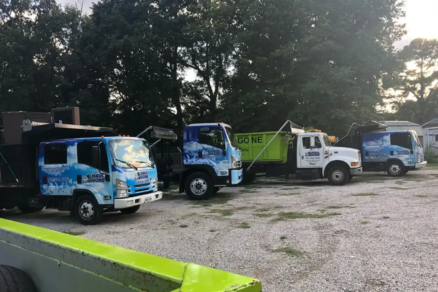 Line of blue and white utility trucks parked on gravel under trees.