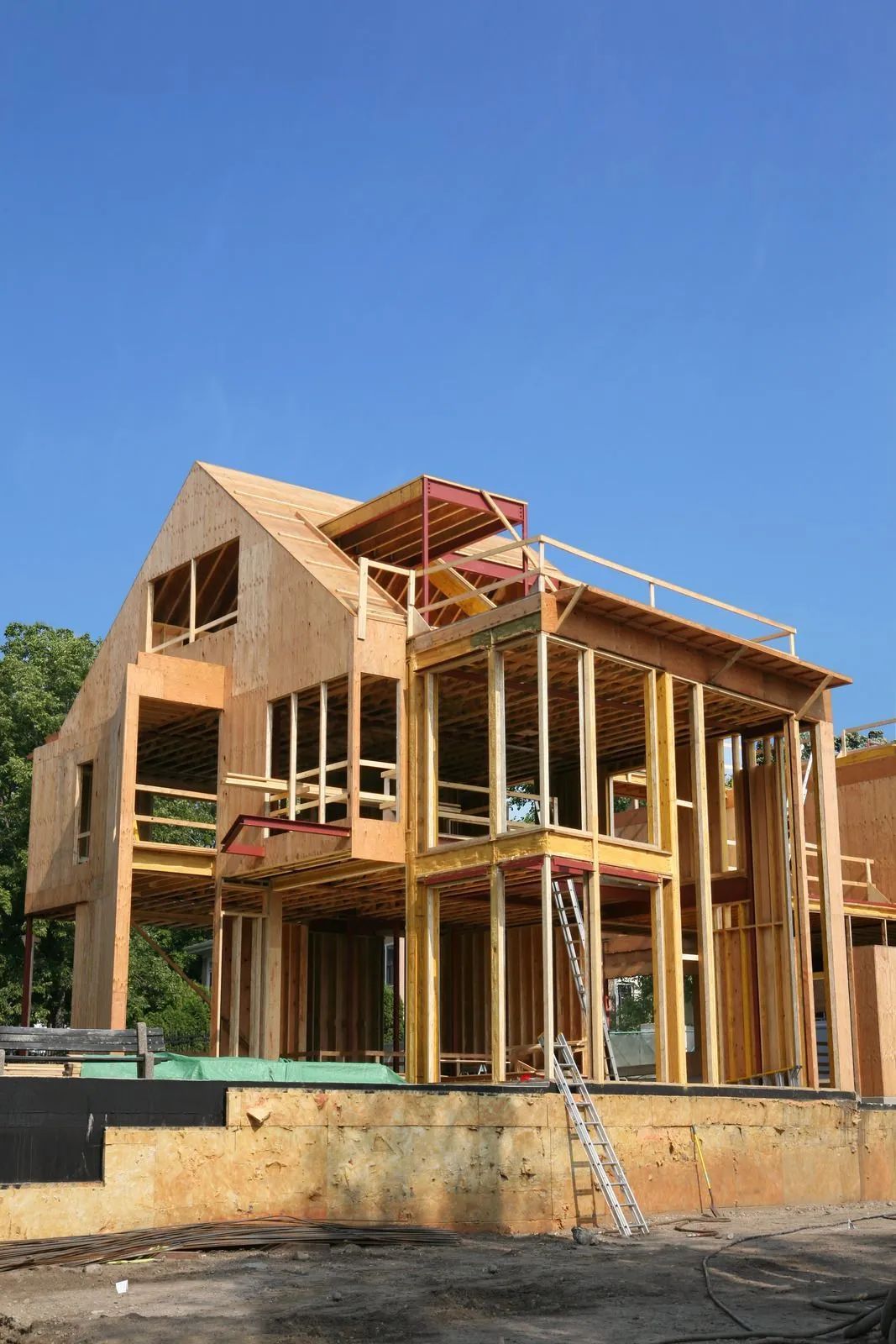 Wood-framed house under construction with scaffolding and a ladder against a blue sky