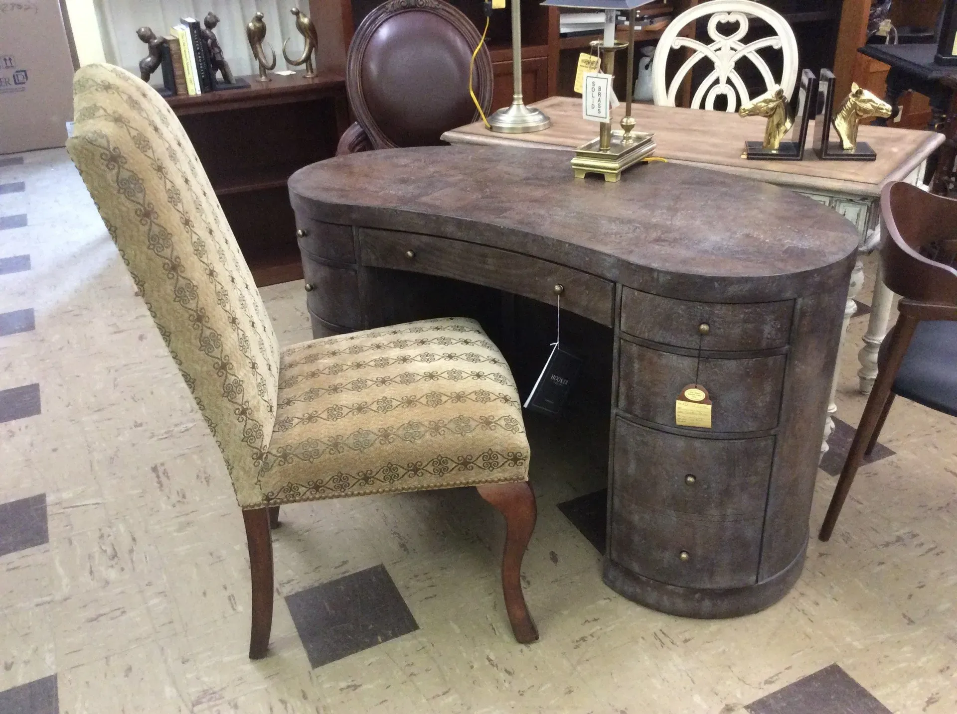 A dark, oval-shaped desk with drawers and a high-backed, patterned upholstered chair in a furniture store.