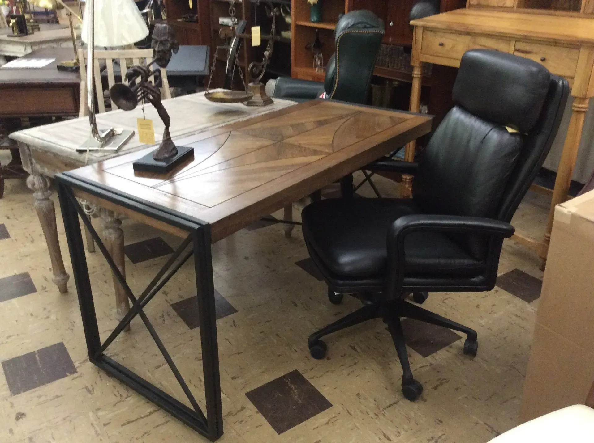 A wooden desk with a black metal X-frame base and a matching black leather rolling office chair in a store setting.