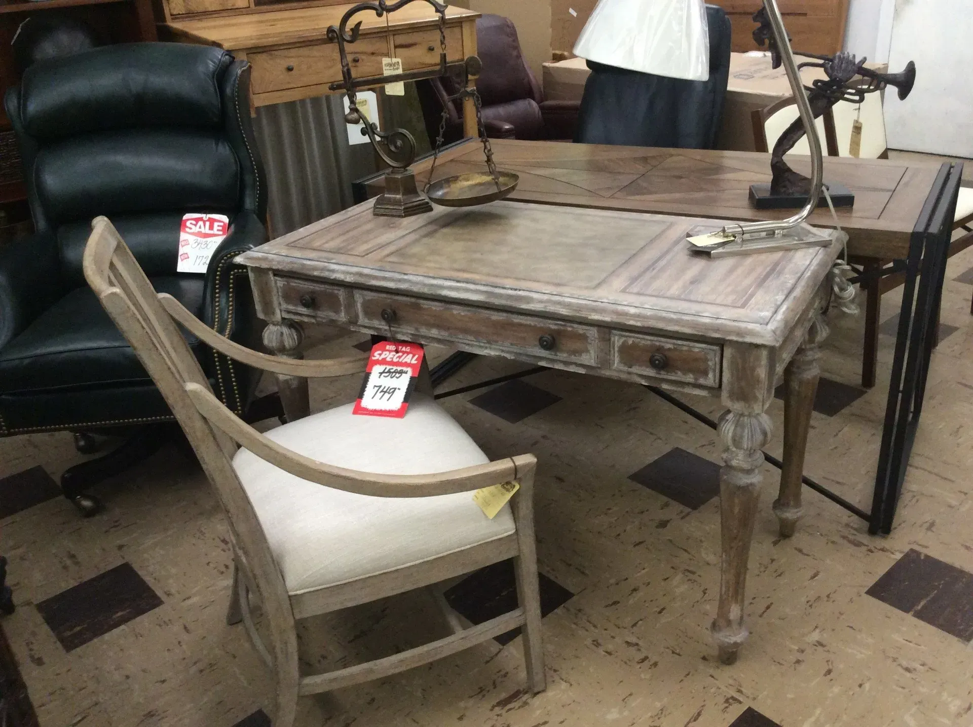 A light-wood desk with three drawers and a matching upholstered chair sitting in a showroom with other office furniture.