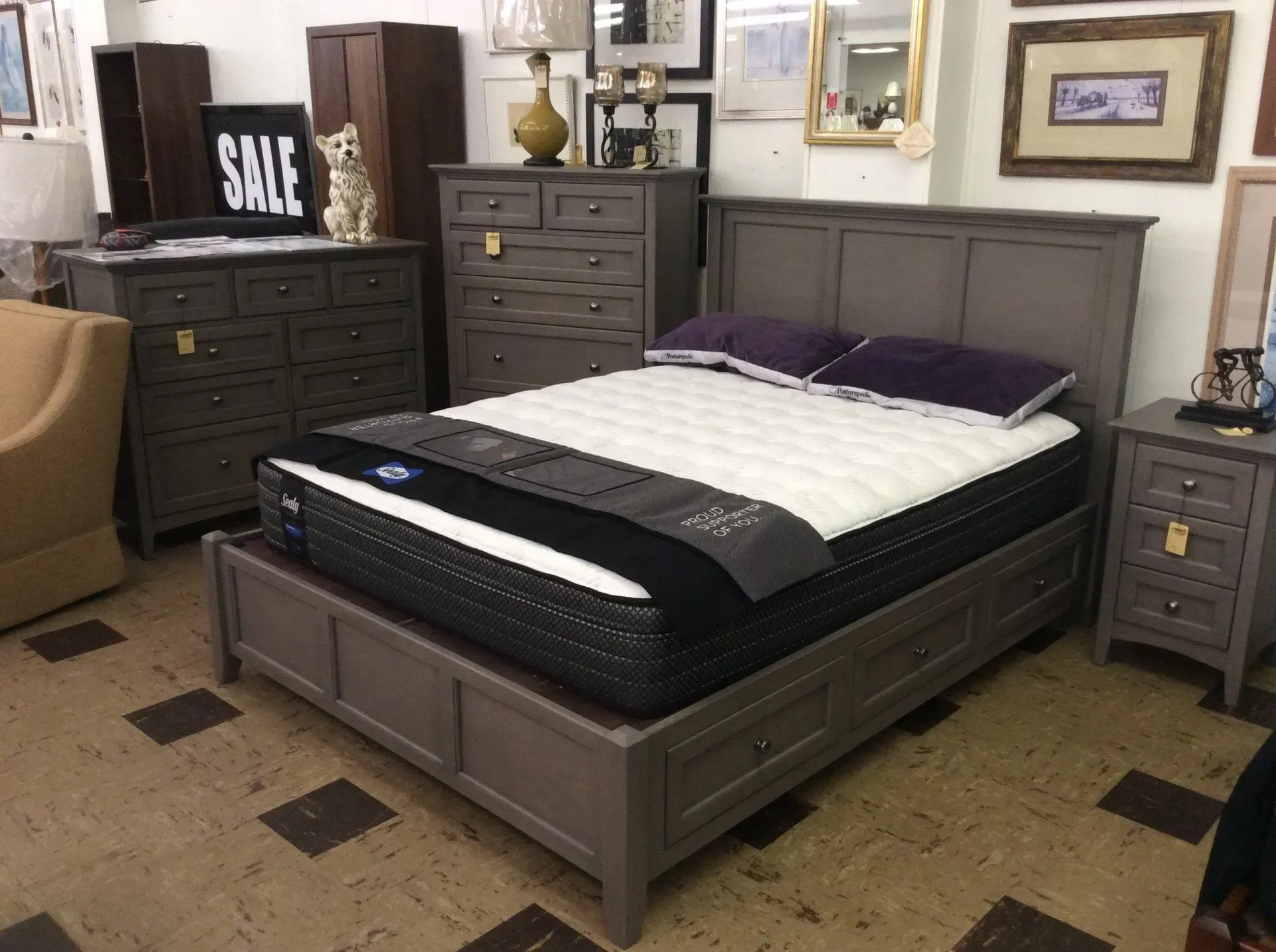 A bedroom set with a grey wood bed, matching dresser, chest of drawers, and nightstand on display in a furniture store.