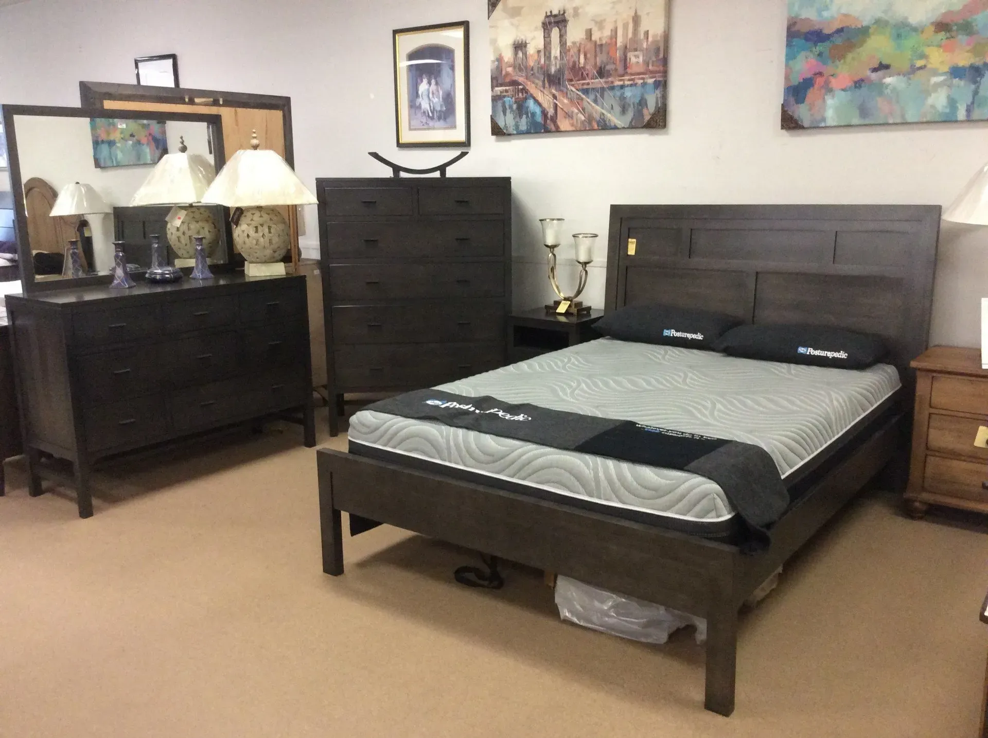 A showroom display featuring a dark wood bedroom set, including a bed with a grey mattress, dresser with mirror, and chest.