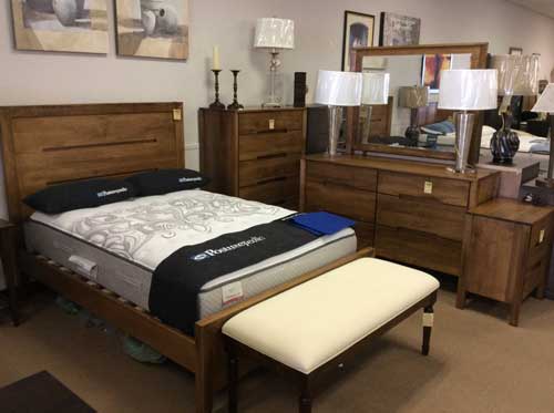 A showroom display featuring a wooden bed frame, matching dresser with mirror, nightstand, and a cream upholstered bench.