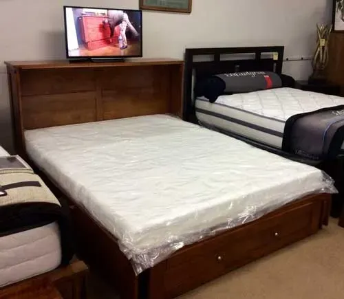A wood storage bed frame with a mattress in plastic, beside another bed, with a flat-screen TV on the wooden headboard.