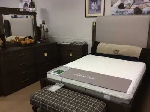 A bedroom display with a dark wood dresser, a beige upholstered headboard, a mattress, and a plaid bench.