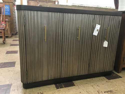 A modern sideboard with vertical, textured gray door panels, dark brown trim, and slim gold vertical handles in a store.