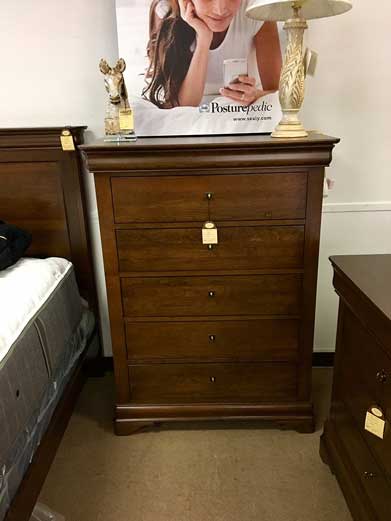 A tall, dark wood dresser with five drawers stands in a showroom next to a bed frame and a lamp.
