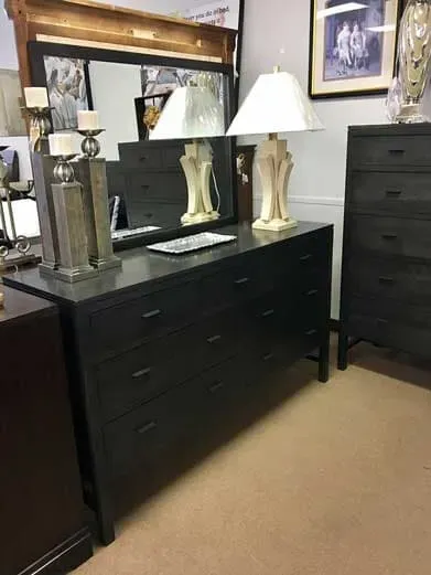 A dark wood dresser with a mirror, two lamps, and a tall chest of drawers in a furniture store.