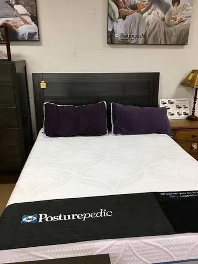 A dark wood bed frame with a white Posturepedic mattress and two purple pillows, set against a wall with framed art.
