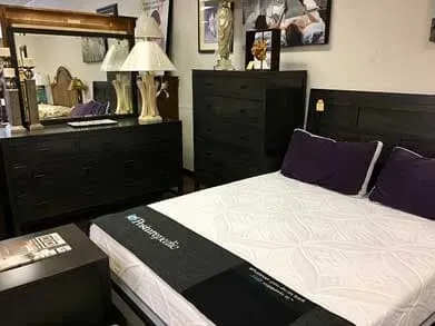 A bedroom display featuring a dark wooden bed frame with purple pillows, a matching dresser with a mirror, and chest.