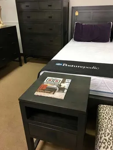 Dark bedroom furniture set including a nightstand, tall dresser, and bed with a white mattress and purple pillow.