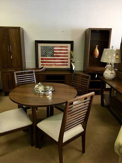 A dining set with a round table and four chairs, arranged in a furniture showroom with wooden cabinets and US flag art.