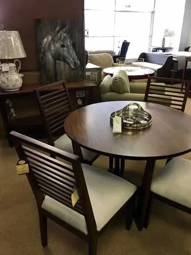 A wooden dining set with a round table and slat-back chairs with light seats in a furniture showroom.