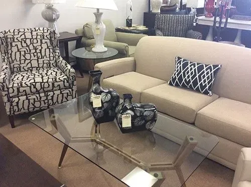 A furniture showroom displays a patterned armchair, a beige sofa with a geometric pillow, and a glass-topped coffee table.