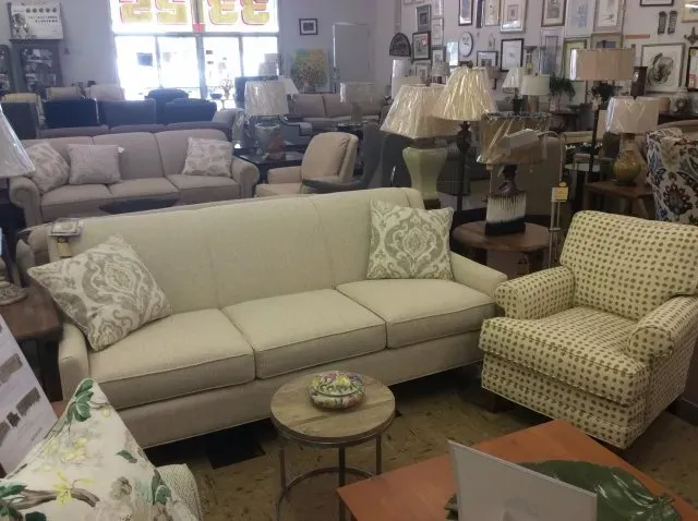 A showroom interior displays a beige sofa with patterned throw pillows, a matching armchair, and various side tables.