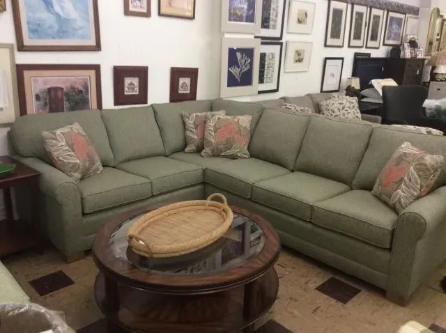 Sage green sectional sofa with patterned accent pillows and a wooden coffee table with a woven tray in a furniture store.