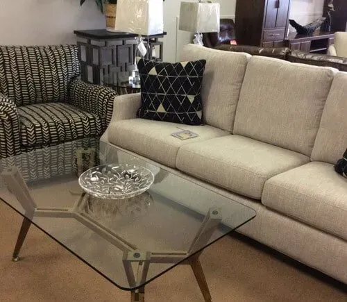 A showroom display featuring a cream-colored sofa with a geometric black pillow, a patterned armchair, and a glass table.