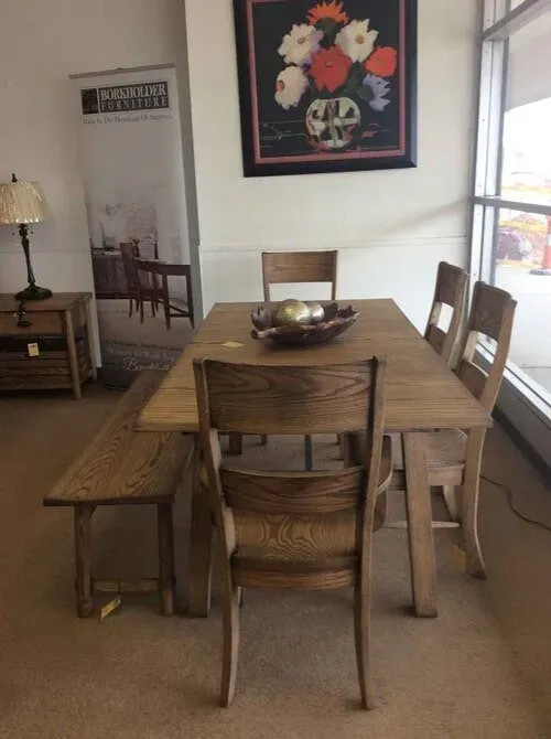 A wooden dining set with a rectangular table, bench, and three chairs, placed in a furniture store beneath a floral painting.