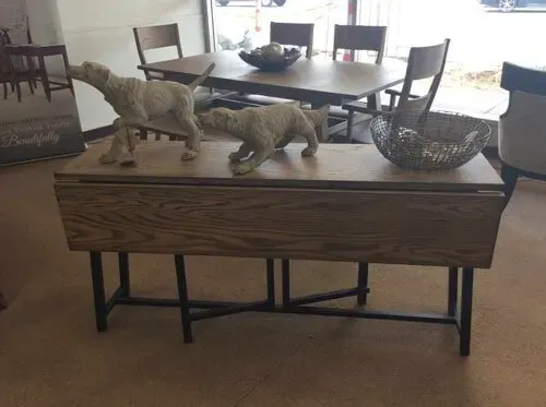 A wooden drop-leaf table with metal legs and two dog statues on top, with a dining set in the background.
