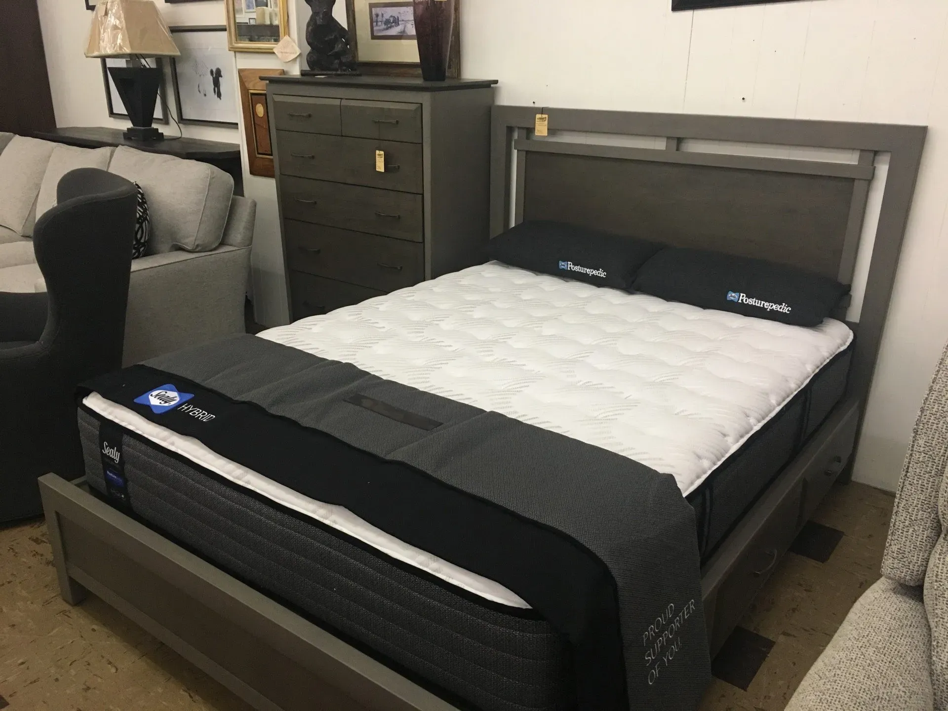 A grey wooden bed frame with a white mattress and black bedding, set in a furniture store next to a matching chest.
