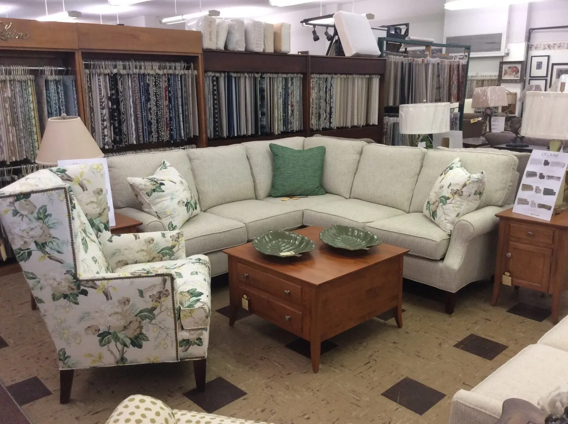 A furniture showroom display featuring an L-shaped beige sectional sofa, a floral wingback chair, and a wooden coffee table.