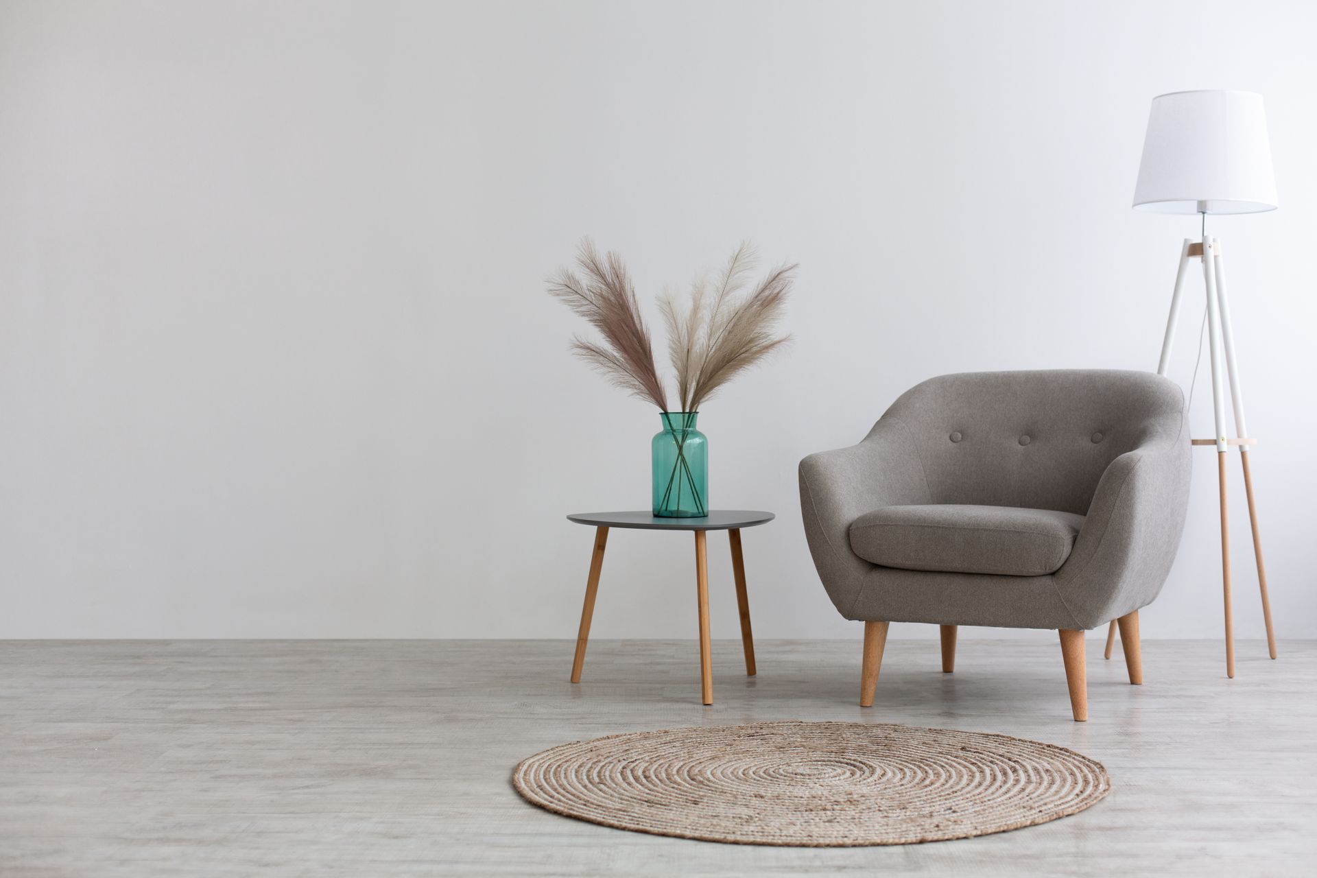A gray armchair, a small table with dried grass in a green vase, and a floor lamp on a circular rug in a minimalist room.