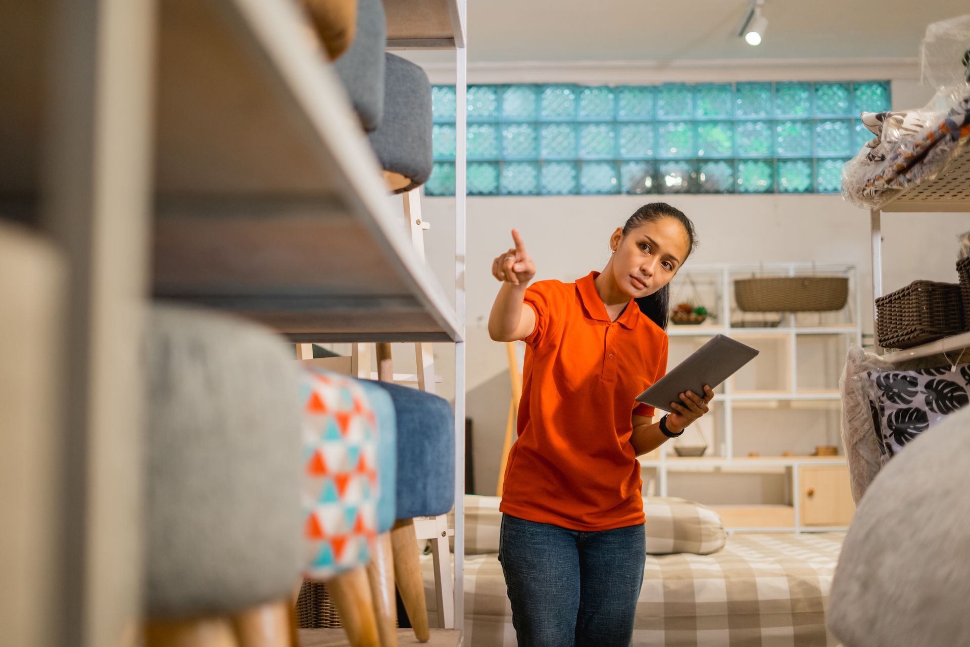 A person in an orange polo shirt holding a tablet points to furniture on shelves in a home goods store.