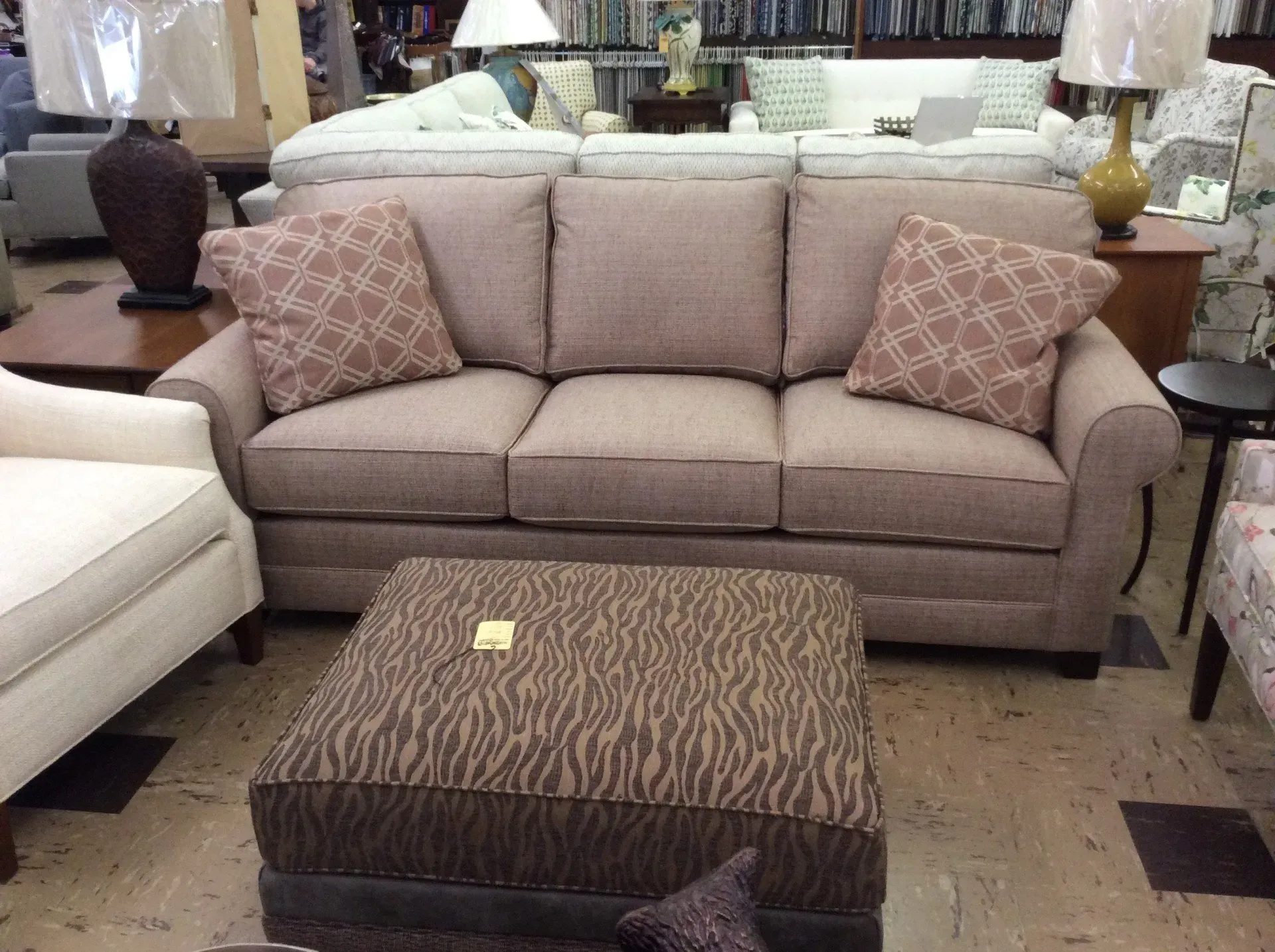 A tan three-cushion sofa with patterned accent pillows and a brown zebra-print ottoman in a furniture showroom.