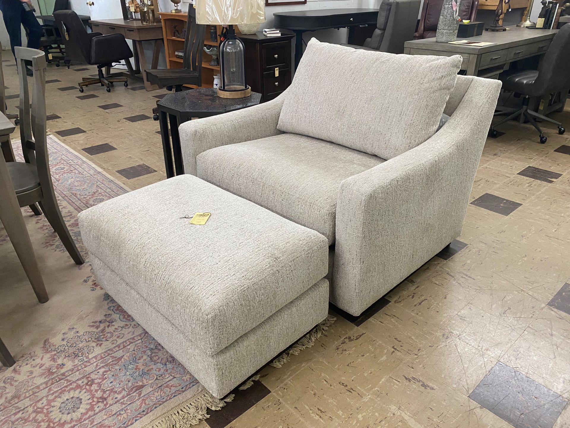 An oatmeal-colored upholstered armchair with a matching ottoman sitting on a patterned rug in a furniture showroom.